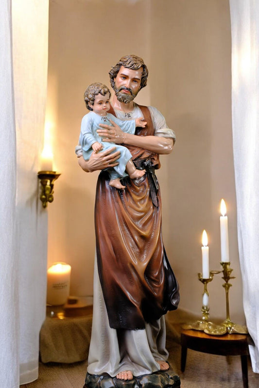 May include: A religious statue depicting a man holding a child. The man wears a brown and white outfit with a beard. The child is dressed in blue. The statue is set in a room with candles.